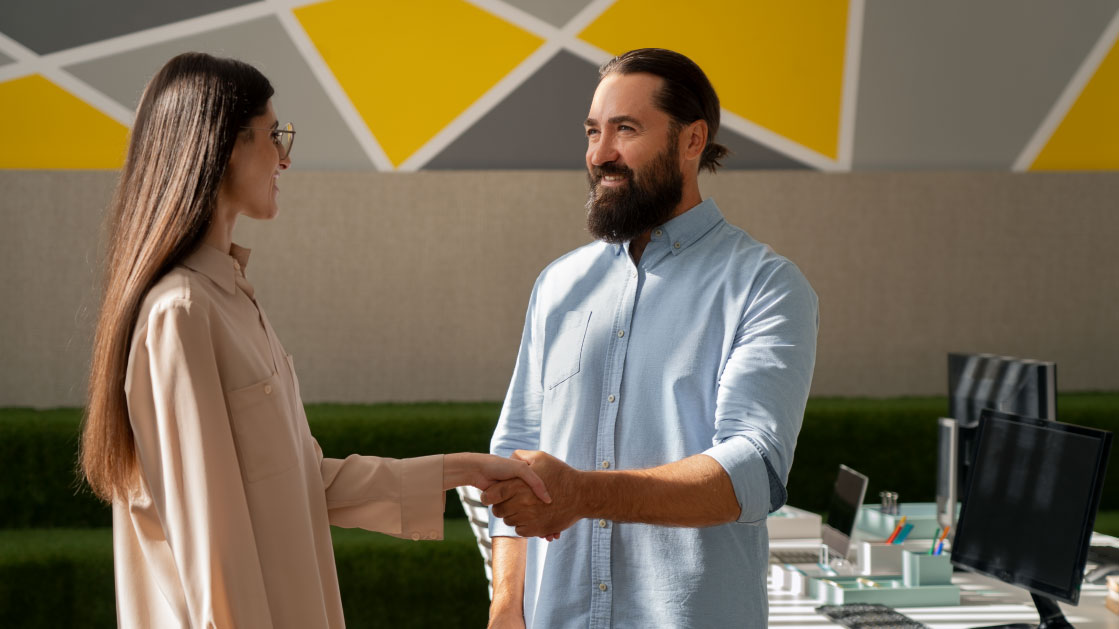 A man in a light blue shirt shakes the hand of a woman in a light pink shirt inside an office.