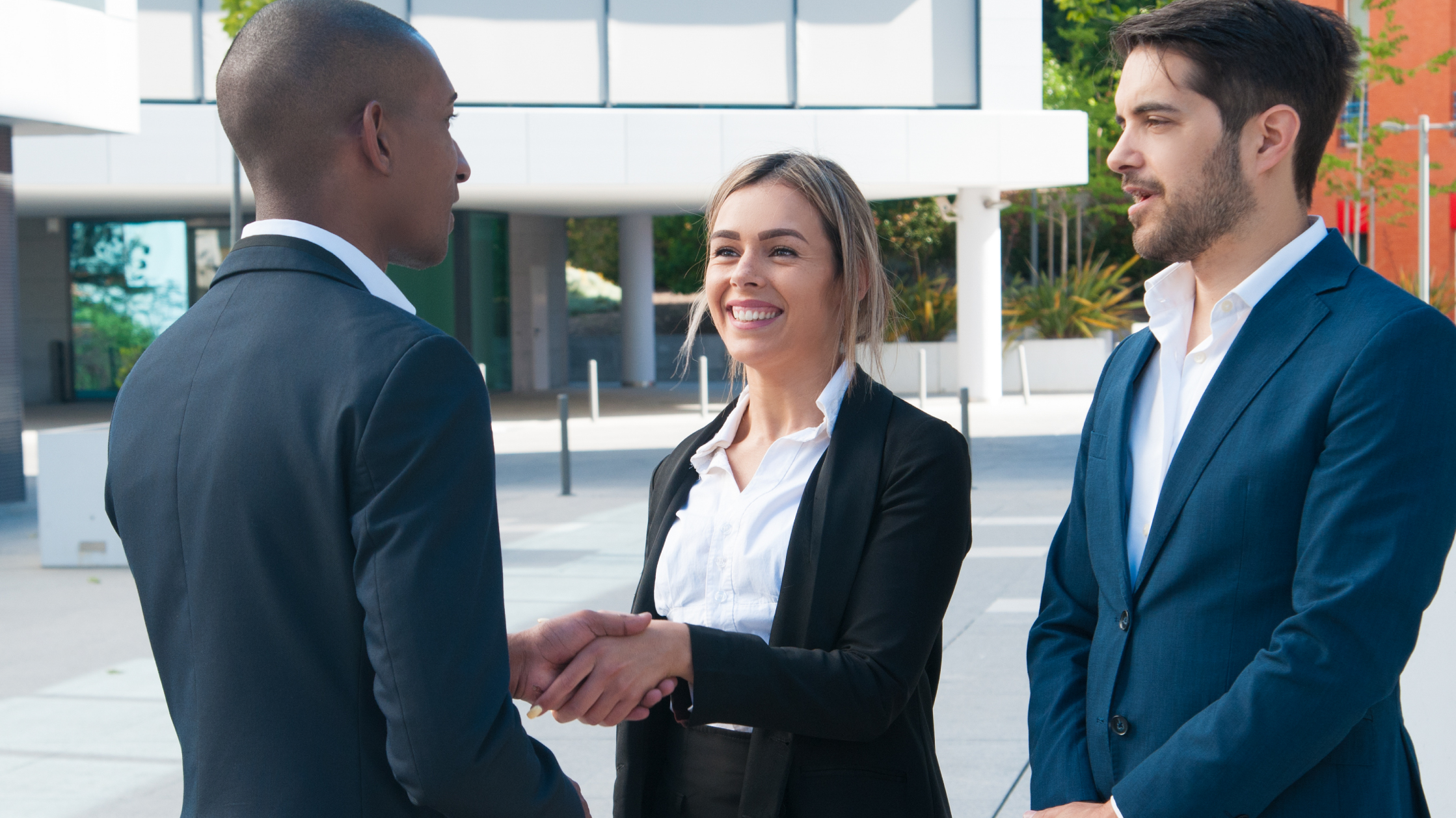 A woman and a man shake hands while a third man watches, They are all wearing suits and white shirts while standing outside a white office building.