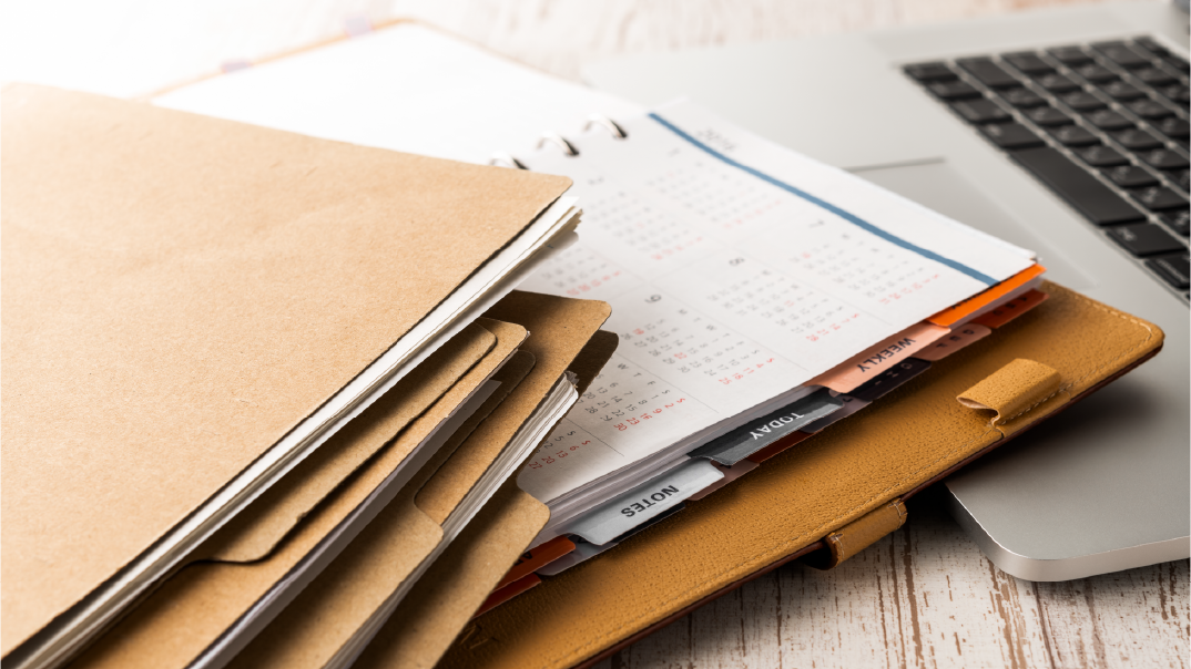 Several manila folders and a yellow agenda laying on top of a grey laptop.