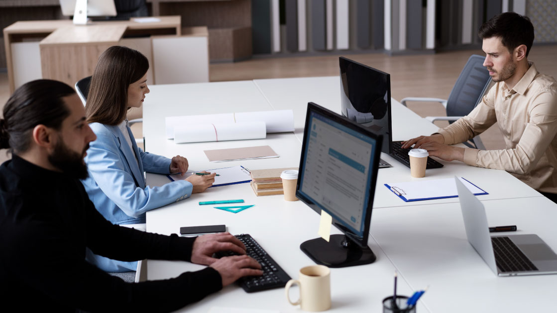A group of two men and a woman work in an office. The men are working on monitors while the woman in a light blue jacket writes on a notepad.