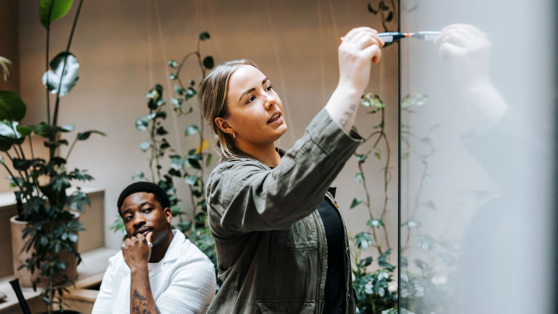 A woman writes on a whiteboard as a man watches behind her. There are a lot of plants behind them.