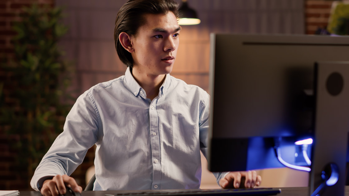 A man in a blue shirt and slicked back black hair works on a monitor inside an office. Behind him is a plant and a lamp.