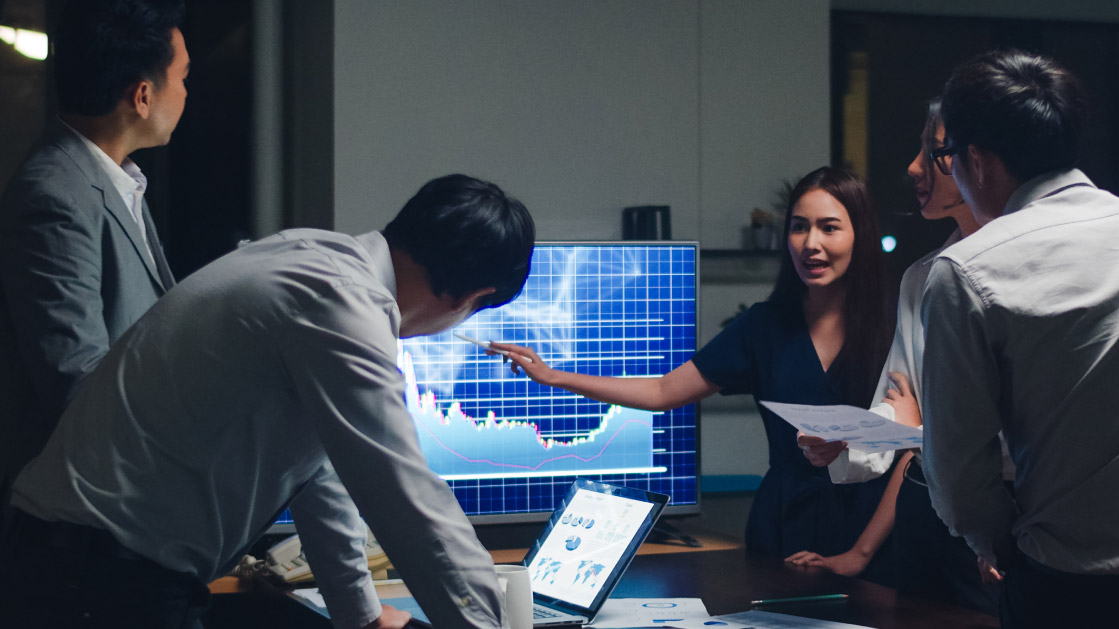 A team of men look at a woman as she points at a graph in a monitor. They are all in a darkened office setting.