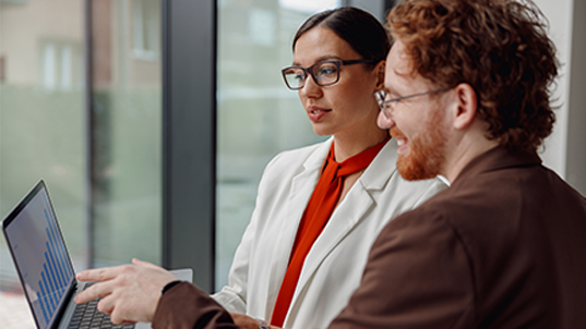 Two people look and discuss a blue graph chart on a laptop. One of them is wearing a white suit jacket and red blouse and the other a dark brown suit. Both of them are wearing glasses.
