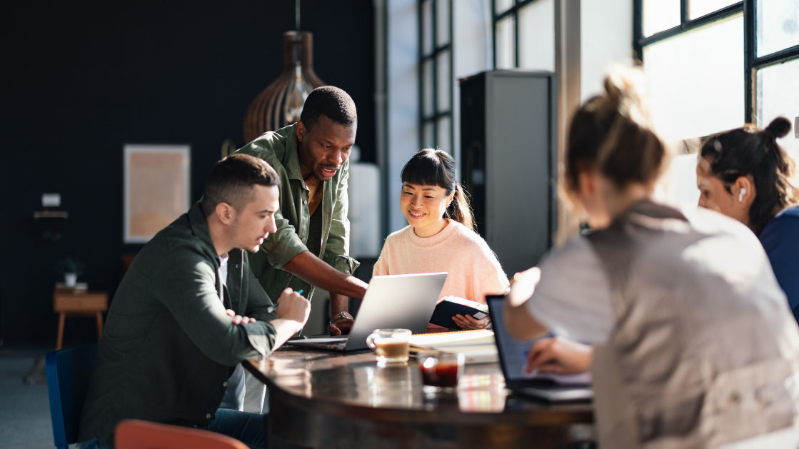 A group of people have a meeting around a table. There are laptops and coffee cups on the table. A man leans over and points to a laptop as another man and a woman lok into it.