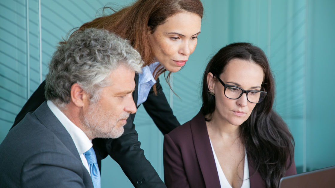 A woman leans in between another woman and a man to look into a computer. 