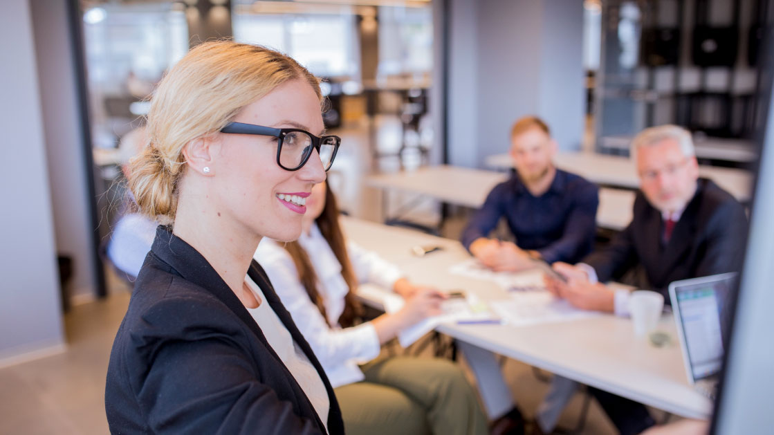 How to Convince your Board to adopt Recruitment Process Outsourcing (RPO): A blonde woman with dark glasses presents to a couple of men on a meeting room in an office.