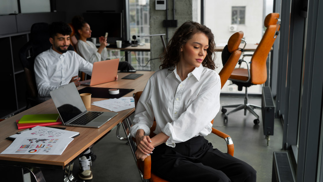 Modern office setting with three people in white shirts and dark pants working at desks equipped with laptops, charts, notebooks, and coffee cups. The room is well-lit by large windows and features orange office chairs.