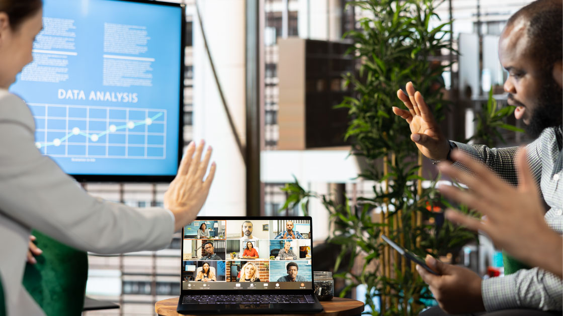 A man and a woman sitting down on a meeting room wave at their team on a video call on a laptop on the center of the image.