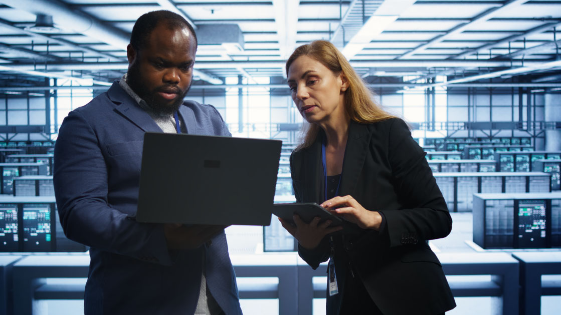 A man and a woman stand inside a server room and look at at laptop.