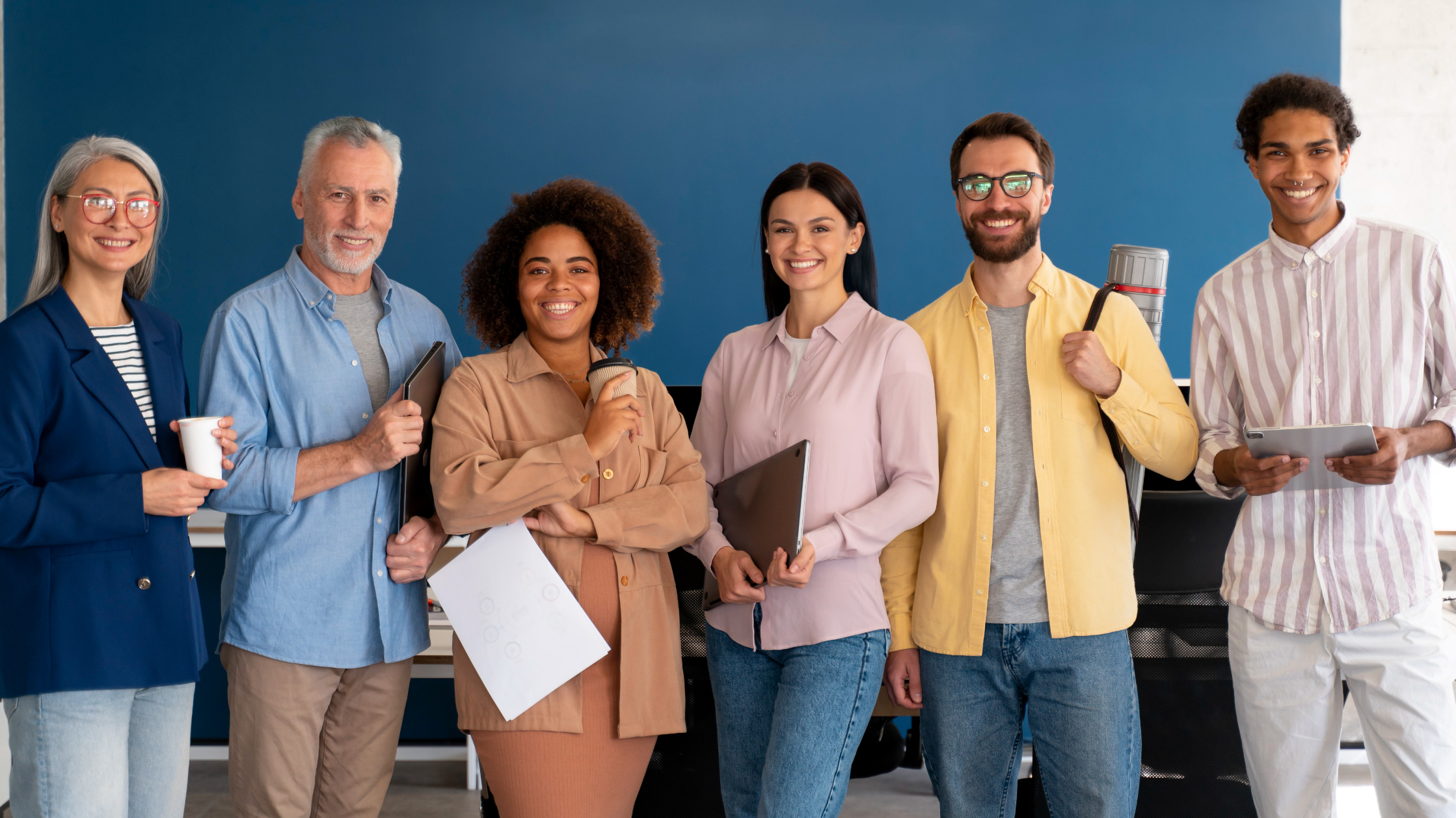 A group of people of different ages and dressed in different colors all smile while holding different objects: paper, a notebook, a coffee cup, and binders.
