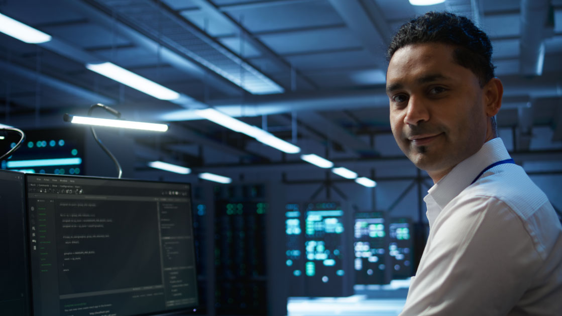 A man on a white t-shirt on a server room smiles as he sits in front of a computer with some HTML code on it.