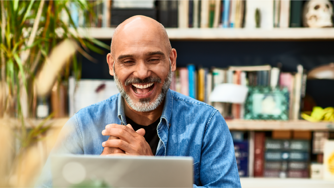 Ace your interview with our guide for job seekers: A man smiles and claps as he looks into his laptop with a bookshelf behind him.