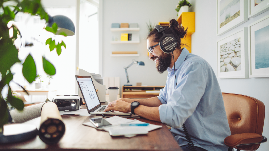 A man smiles as he writes his resume on his laptop. He is wearing headphones and working in home office in his living room.