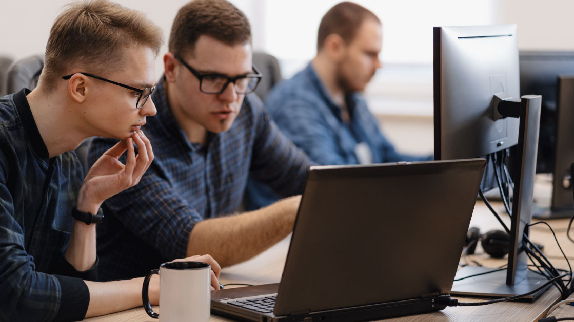 Three men in blue plaid shirts work on monitors and laptops. Two of them are wearing black-rimmed glasses while looking engaged in their work.