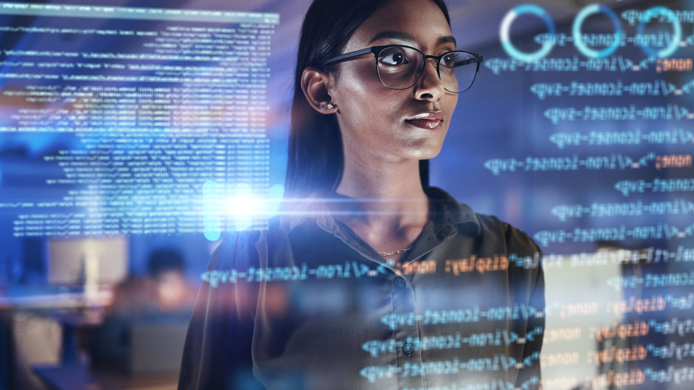 A woman wearing a grey shirt and black glasses looks at several lines and windows of code and data in front of her.
