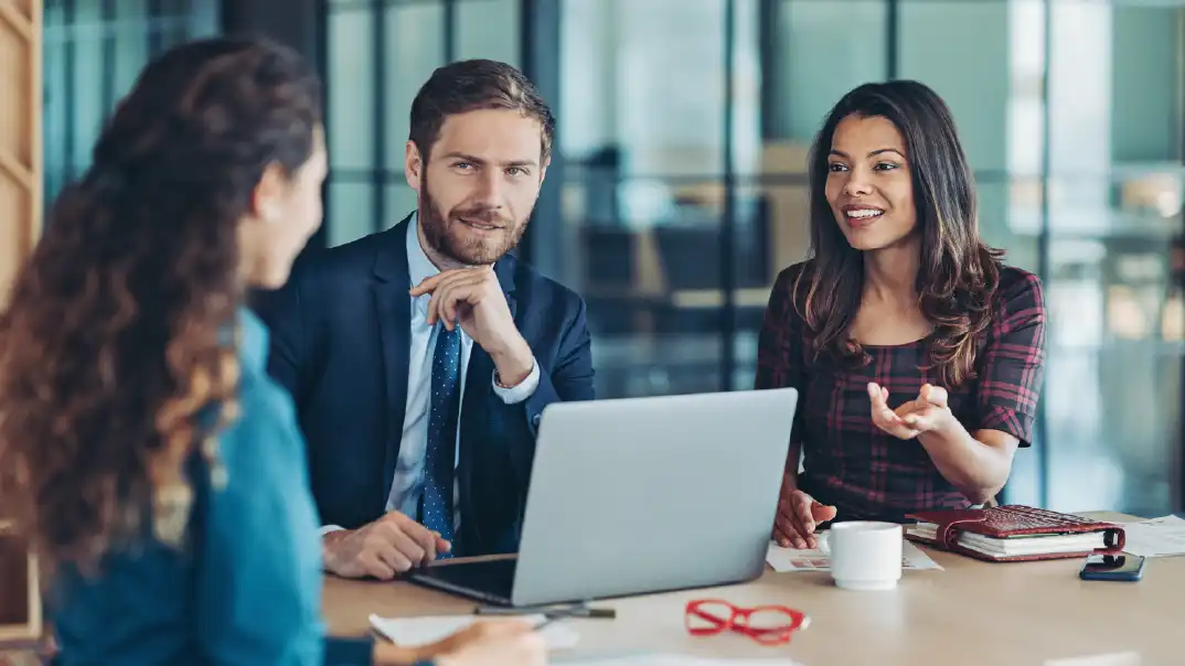 Three professionals meeting at a table with a laptop, discussing workforce strategy in a modern office.