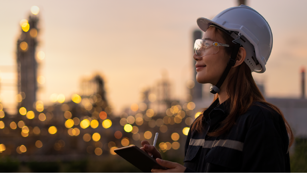 A woman superintendent looks ahead. She is wearing a white hard hat and dark safety bet. On the background and unfocused is a city with bright yellow lights.