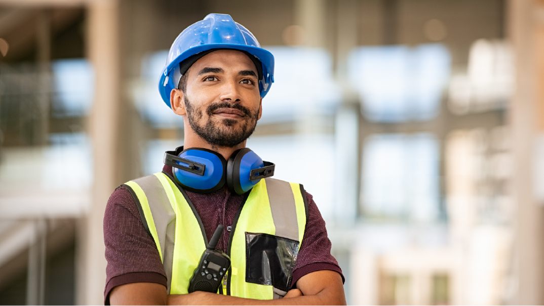 A man in a construction site looks on with satisfaction. He is wearing a red shirt, blue hard hat and blue protective headphones, and a bright yellow safety vest with a black radio.