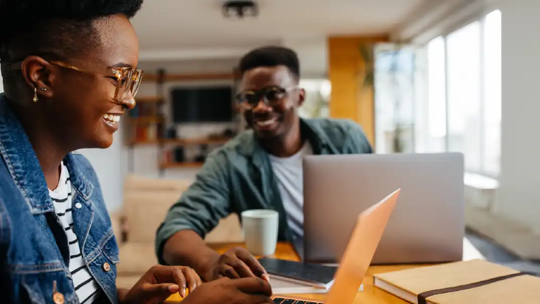 Two people work together at home. They're both on their laptops and smiling, having a good time. They are also both wearing glasses.