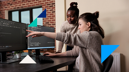 Two women look at double monitors displaying a coding program on an office. One of them is pointing at something on the screen.