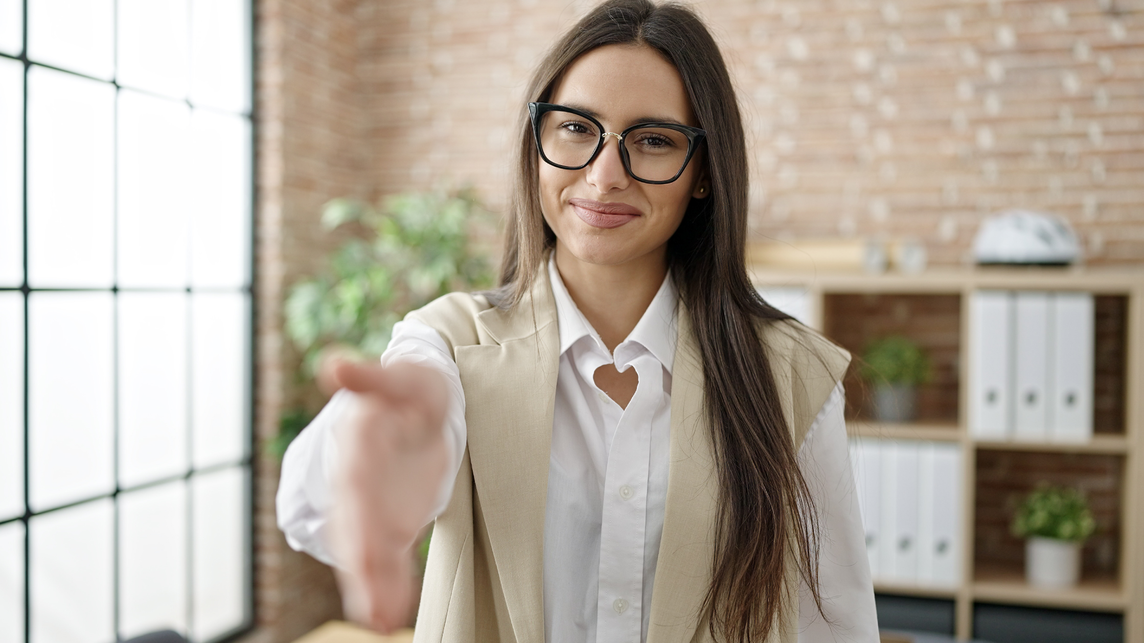 A woman in a white shirt and beige vest extends her hand as if to shake it. She has long, brown and straight hair and black glasses.