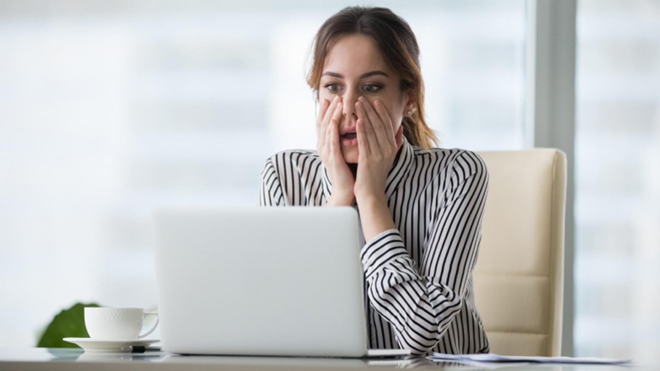 A woman in an office looks at her laptop and holds her hands to her face in an astonished expression like if she has made a mistake. There's a coffee cup to her left.