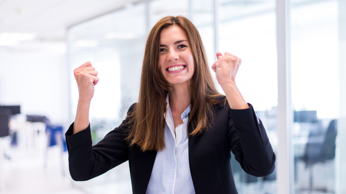 A woman with raised fists and a big smile in her face. She has long brown straight hair, a white shirt and a black suit jacket.
