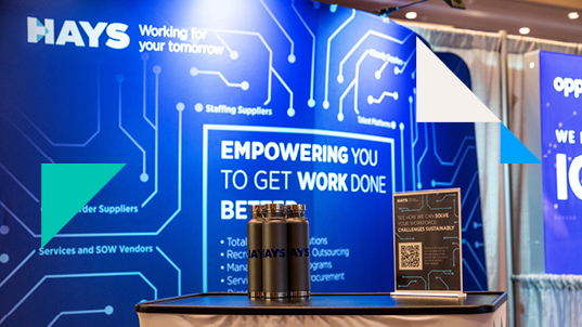 On the background there's a banner with the words: Empowering you to get work done better. In front of it there's a table with grey water bottles with the Hays logo and a paper stand with a QR code.
