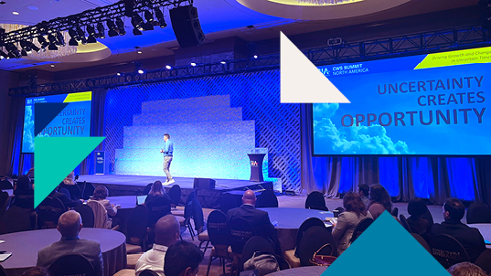 Blue-tinted image of a man giving a conference in front of an audience. He stands between two screens with the text: Uncertainty creates opportunity.