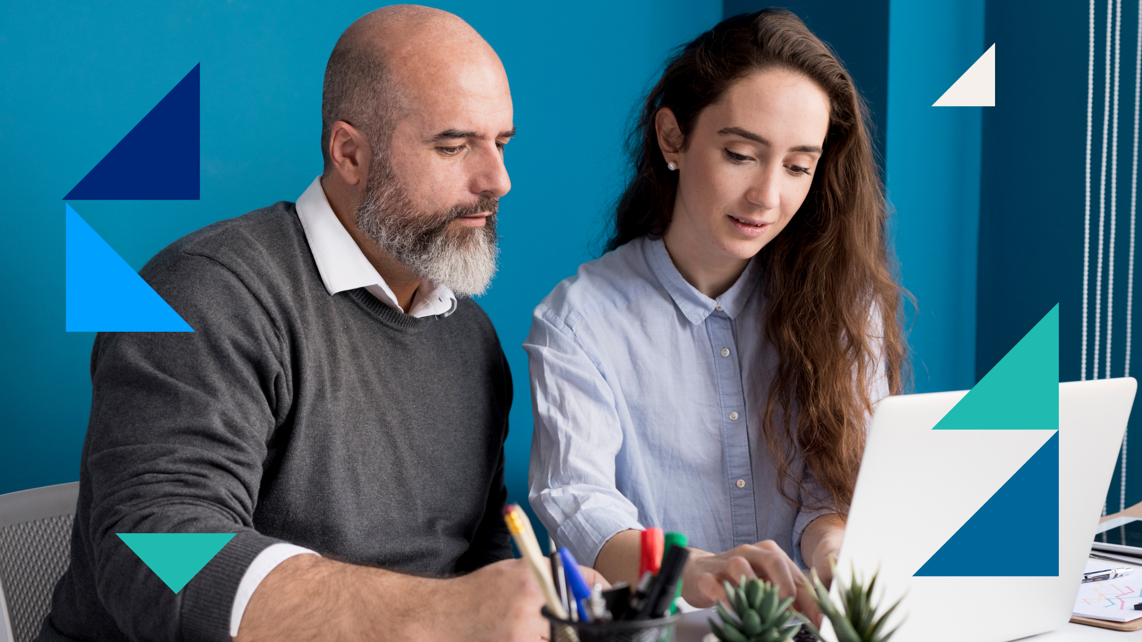 A man and a woman look at a laptop, collaborating together. You can see a some pens and plants on the desk. The background is bright blue.