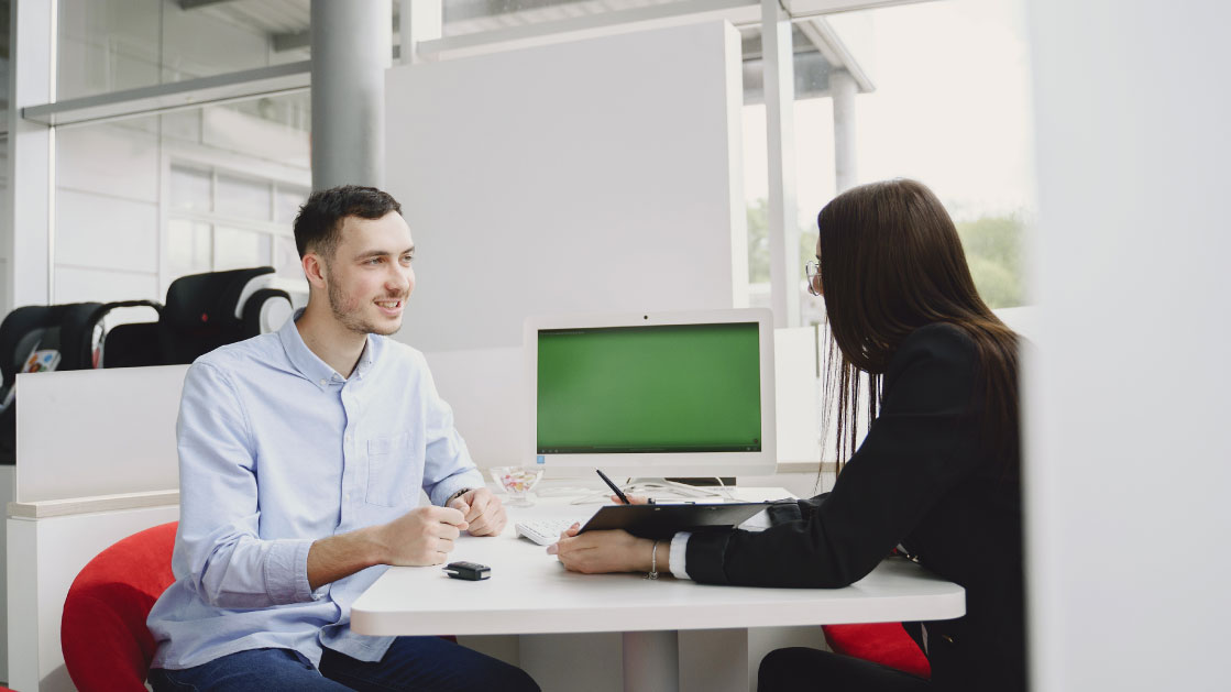 A man in a blue shirt and a woman in a dark suit sit in front of the other and talk in a desk with a white computer on the backgorund. They seem to be in a white office.