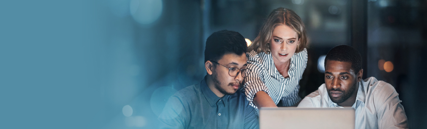 Three people collaborating around a laptop inside a dark office with a blurred background.