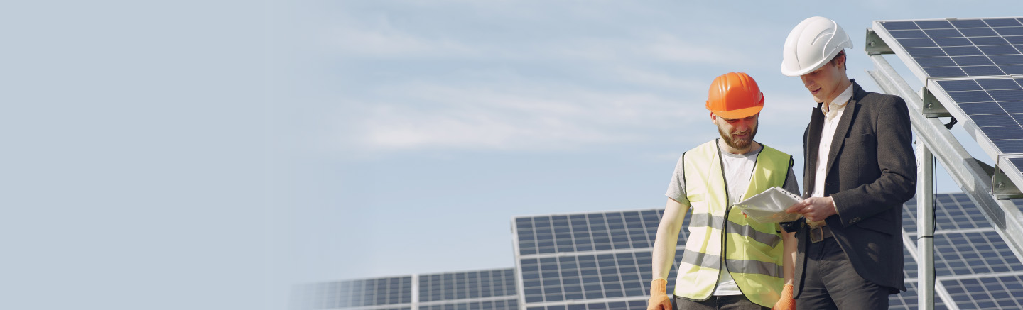 Two men look into a clipboard. One of them is using a suit and a white hard hat, the other wears protective gear including a yellow vest, orange vest and globes. Behaind them there's a series a solar panels.