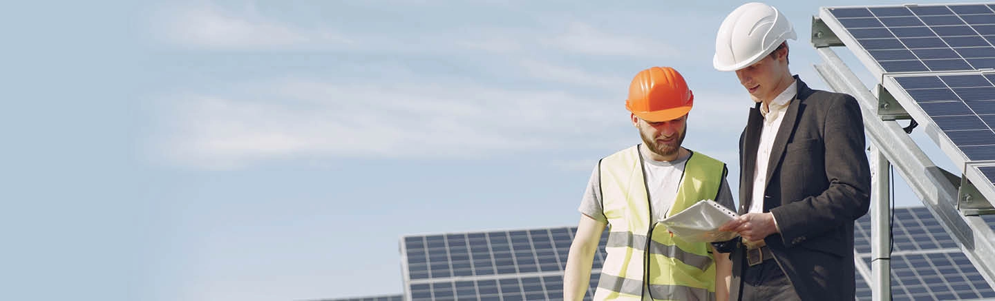 Two construction workers look into a folder. One of them is wearing a black suit jacket and white shirt and white hardhat, the other wears a bright orange hard hat and a bright yellow construction vest. Behind them are rows of solar panels.