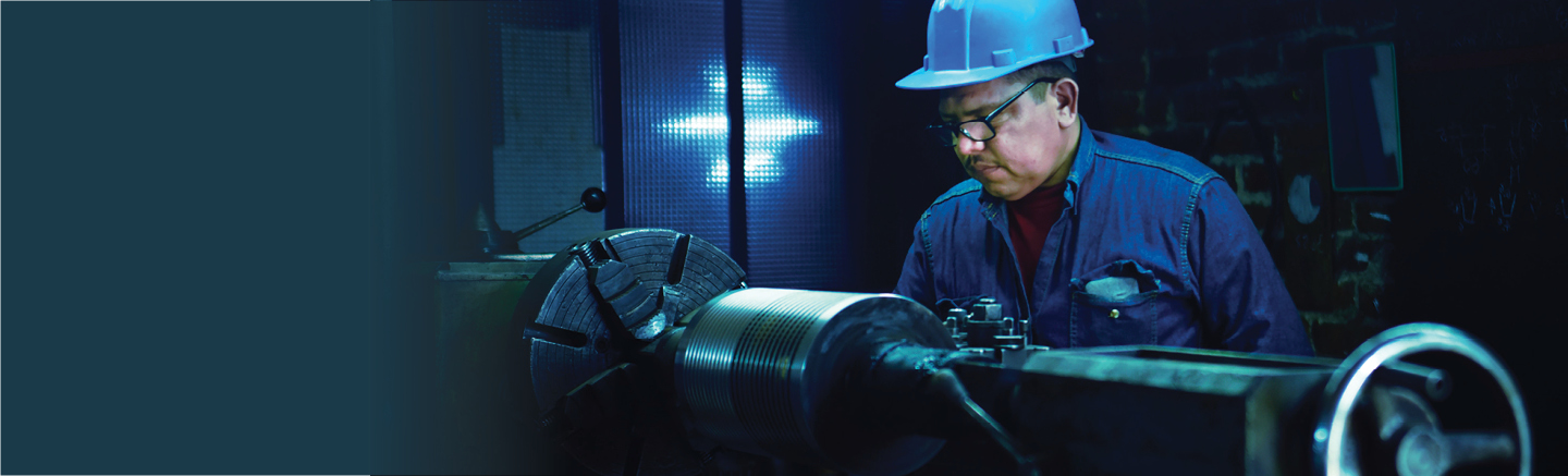 Hays are the experts in manufacturing staffing across the United States. A man in a white hardhat and glasses works on machinery. He is wearing a blue denim shirt and underneath a red shirt.