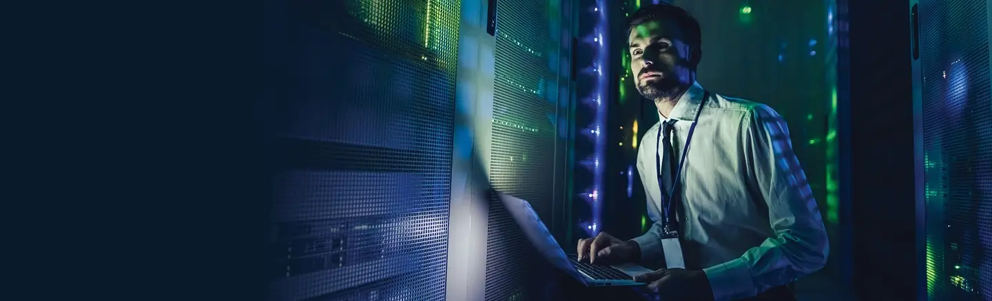 Person in white shirt and tie working on a laptop in a server room with lit server racks.