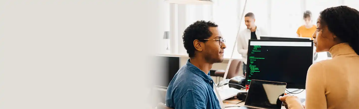 A group of people in an office setting focused on software development. Two individuals sit at a desk with monitors displaying code, one holds a laptop, and another stands in the background. Faces are blurred for privacy.
