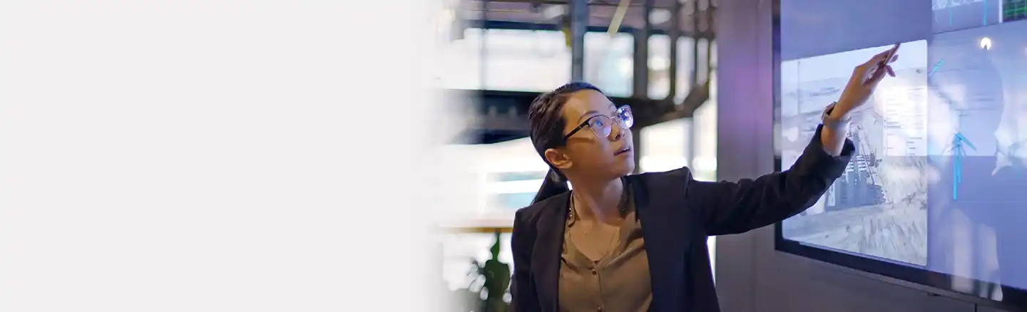A person in a business suit stands in front of a large screen displaying charts and graphs, pointing at the screen. The office setting features large windows and modern decor, with the person's face pixelated for privacy